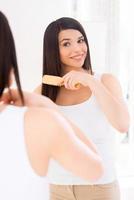 donna pettinatura capelli. attraente giovane donna pettinatura sua capelli mentre guardare a il specchio foto