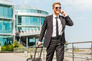 esecutivo su il andare. bello giovane uomo d'affari nel pieno completo da uomo trasporto valigia e parlando su il mobile Telefono mentre a piedi all'aperto foto