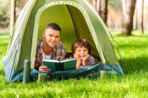 campeggio insieme è divertimento. padre e figlio lettura libro e sorridente mentre dire bugie nel tenda insieme foto