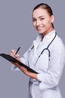 contento femmina medico. fiducioso femmina medico nel bianca uniforme scrittura appunti mentre in piedi contro grigio sfondo foto