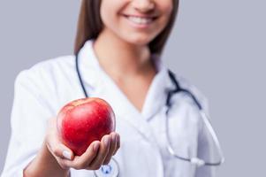 mangiare salutare avvicinamento di femmina medico nel bianca uniforme Tenere rosso Mela e sorridente mentre in piedi contro grigio sfondo foto