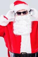 freddo babbo natale. Santa Claus nel occhiali da sole regolazione il suo cuffie mentre in piedi contro grigio sfondo foto