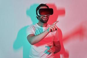 fiducioso giovane uomo nel virtuale la realtà occhiali Esprimere a gesti con colorato ombre nel il sfondo foto