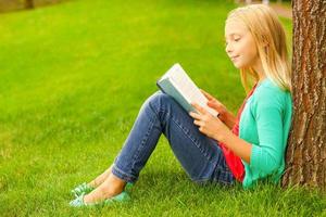 carino poco topo di biblioteca. carino poco biondo capelli ragazza lettura libro mentre seduta su verde erba e pendente a il albero foto