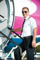 pronto per volo fiducioso maschio pilota nel uniforme andando in il aereo e sorridente foto