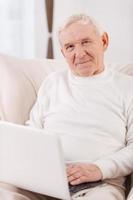 fare surf il rete. fiducioso anziano uomo Lavorando su il computer portatile e guardare a telecamera mentre seduta nel sedia a il suo appartamento foto