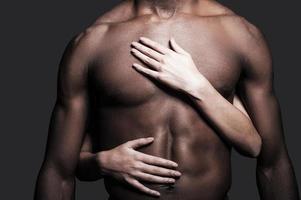 amorevole Questo corpo. avvicinamento di A petto nudo africano uomo con femmina mani Abbracciare una persona il suo torso contro grigio sfondo foto
