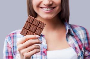 cioccolato umore. avvicinamento di giovane donne Tenere bar di cioccolato e sorridente mentre in piedi contro grigio sfondo foto