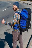 continuando il in viaggio. bello giovane uomo trasporto zaino allungamento su mano e guardare a telecamera attraverso il le spalle con pollice su mentre in piedi su il strada foto