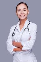 assunzione bene cura di il tuo Salute. fiducioso femmina medico nel bianca uniforme guardare a telecamera e sorridente mentre in piedi contro grigio sfondo foto