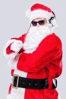 allegro Natale per voi Santa Claus nel occhiali da sole e cuffie puntamento voi mentre in piedi contro grigio sfondo foto