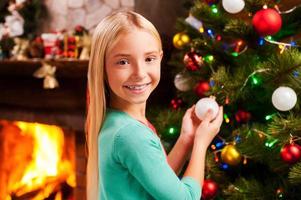 preparazione per Natale. carino poco ragazza decorazione Natale albero e sorridente con camino nel il sfondo