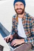 come un' bene giorno per Lavorando all'aperto bello giovane barbuto uomo sorridente mentre Lavorando su il computer portatile e guardare a telecamera foto