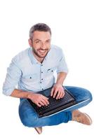 contento utente. superiore Visualizza di bello giovane uomo nel casuale indossare seduta su il pavimento e Lavorando su il computer portatile mentre essere isolato su bianca sfondo foto