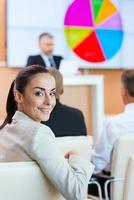 donna d'affari a il conferenza. fiducioso giovane donna nel vestito formale guardare al di sopra di spalla e sorridente mentre seduta a il conferenza sala con pubblico altoparlante nel il sfondo foto