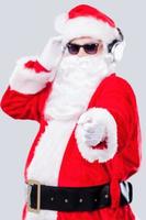 allegro Natale per voi freddo Santa Claus nel occhiali da sole regolazione il suo cuffie e puntamento voi mentre in piedi contro grigio sfondo foto