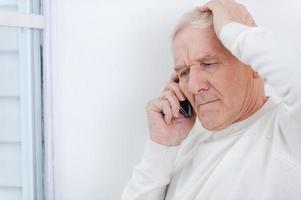 avendo alcuni guai. frustrato anziano uomo parlando su il mobile Telefono e toccante il suo testa mentre pendente a il parete foto