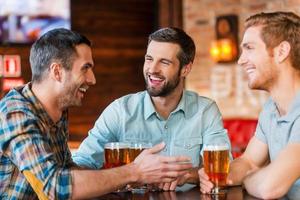 incontro con il migliore gli amici. tre contento giovane uomini nel casuale indossare parlando e potabile birra mentre seduta nel bar insieme foto