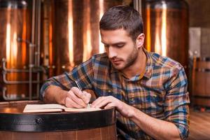 pianificazione il suo birra attività commerciale. fiducioso giovane uomo nel casuale camicia scrittura qualcosa nel il suo Nota tampone mentre pendente a il di legno barile con metallo contenitori nel il sfondo foto