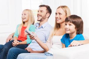 Guardando loro preferito mostrare. lato Visualizza di contento famiglia di quattro bonding per ogni altro e sorridente mentre Guardando tv a casa insieme foto