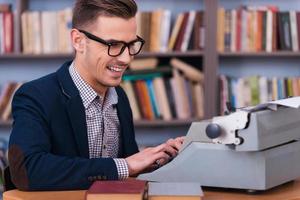 riuscito autore. lato Visualizza di contento giovane autore digitando qualcosa a il macchina da scrivere e sorridente mentre seduta a il suo Lavorando posto con scaffale nel il sfondo foto