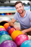 bello e fiducioso giocatore. allegro giovane uomo seduta vicino bowling palle e sorridente contro bowling vicoli foto