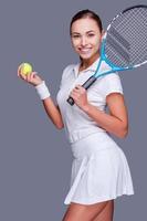 portando il bellezza per il gioco. bellissimo giovane donne nel gli sport Abiti Tenere tennis racchetta su sua spalla e sorridente mentre in piedi contro grigio sfondo foto