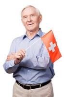svizzero amicizia. allegro anziano uomo Tenere bandiera di Svizzera e sorridente a telecamera mentre in piedi contro bianca sfondo foto