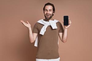 contento giovane uomo Tenere inteligente Telefono e Esprimere a gesti mentre in piedi contro Marrone sfondo foto