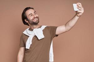 contento giovane uomo fabbricazione autoscatto di inteligente Telefono contro Marrone sfondo foto