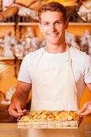 il fresco al forno biscotti per voi. bello giovane uomo nel grembiule Tenere vassoio con biscotti e sorridente mentre in piedi nel forno negozio foto