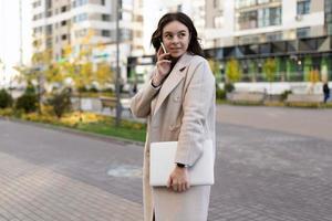 riuscito giovane attività commerciale donna con un' il computer portatile nel sua mani parla su un' mobile Telefono contro il fondale di il città, forte e indipendente donna concetto foto