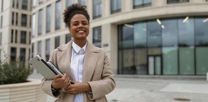 ritratto di un' riuscito giovane africano americano donna con un' il computer portatile nel sua mani e un' largo Sorridi contro il fondale di un ufficio costruzione, concetto di riuscito attività commerciale negoziati foto