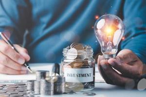 leggero lampadina nel un' di uomo d'affari mano e un' mucchio di monete e monete nel un' bicchiere jar.all su tavolo, soldi Salvataggio e investimento idee e profitto crescita, finanziaria gestione e attività commerciale strategia.per successo foto