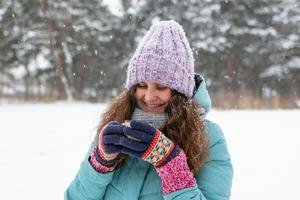 bellissimo sorridente donna con lungo Riccio capelli nel blu inverno giacca e a maglia colorato guanti è Tenere nel sua mani tazza di caldo tè su un' camminare nel inverno nevoso foresta. foto