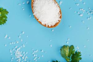 grande bianca mare sale nel un' di legno cucchiaio su un' blu sfondo, con sale e erbe aromatiche sparpagliato in giro il bordi. foto