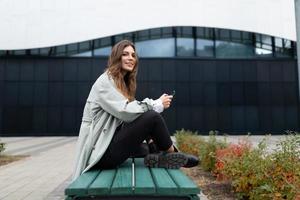 un' giovane donna si siede su un' panchina a gambe incrociate sotto sua con un' mobile Telefono guardare a il telecamera su il sfondo di il città foto