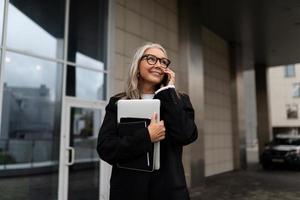 un' attività commerciale anziano donna parla su un' mobile Telefono con un' il computer portatile nel sua mani contro il fondale di un ufficio edificio foto