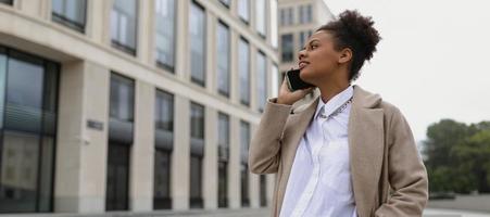 giovane riuscito donna assicurazione agente di africano americano aspetto parla su un' mobile Telefono contro il sfondo di un ufficio edificio foto