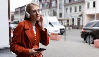 un' giovane donna ascolta per musica nel cuffie con un' Telefono nel sua mani contro il fondale di città caffè foto