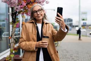 elegante anziano donna comunica di mobile Telefono video chiamata nel il città con un' tazza di caffè nel sua mani foto