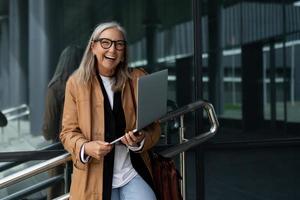 cinquantenne riuscito donna d'affari con un' il computer portatile nel sua mani sorrisi a grandi linee al di fuori il ufficio foto
