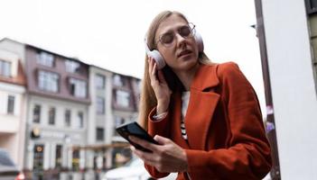 giovane bionda donna con un' mobile Telefono nel sua mani ascolta per musica su cuffie contro il fondale di il urbano paesaggio