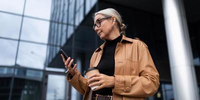 elegante anziano donna con un' mobile Telefono e un' bicchiere di caffè su il sfondo di un' moderno attività commerciale centro, carriera crescita concetto foto