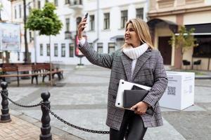 elegante attività commerciale adulto donna con un' il computer portatile nel sua mani prende un' autoscatto su un' mobile Telefono vicino il città piazza foto