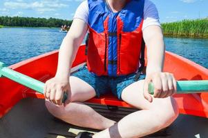 un' uomo nel un' rosso barca, pantaloncini e un' vita giacca è paddling con remi su un' barca per un' camminare su il acqua di il lago fiume mare nel natura foto