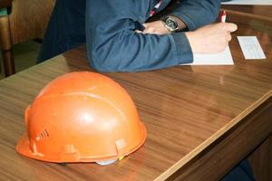 un' uomo Lavorando come un ingegnere con un arancia giallo casco su il tavolo è studiando, scrittura nel un' taccuino a un industriale pianta fabbrica foto