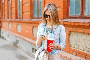 bellissimo donna nel un' denim giacca Tenere un' rosso tazza e scrive un' Messaggio su il Telefono su il sfondo di un' mattone costruzione. foto