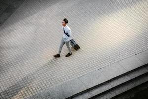 contento giovane uomo d'affari a piedi con valigia nel il città. moderno maschio nel attività commerciale viaggio. alto angolo Visualizza. pieno lunghezza foto