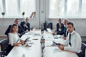 gruppo di persone nel elegante vestito formale guardare a telecamera e utilizzando moderno tecnologie mentre Lavorando nel il ufficio foto